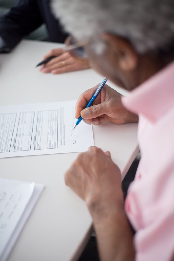 Close-up of a senior adult signing a document with a blue pen on a white table.
